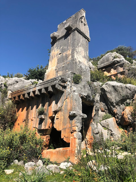 The monumental giant Lycian sarcophagus rising among the greenery at the entrance of Sura Ancient City, with the rugged acropolis hill in the background.