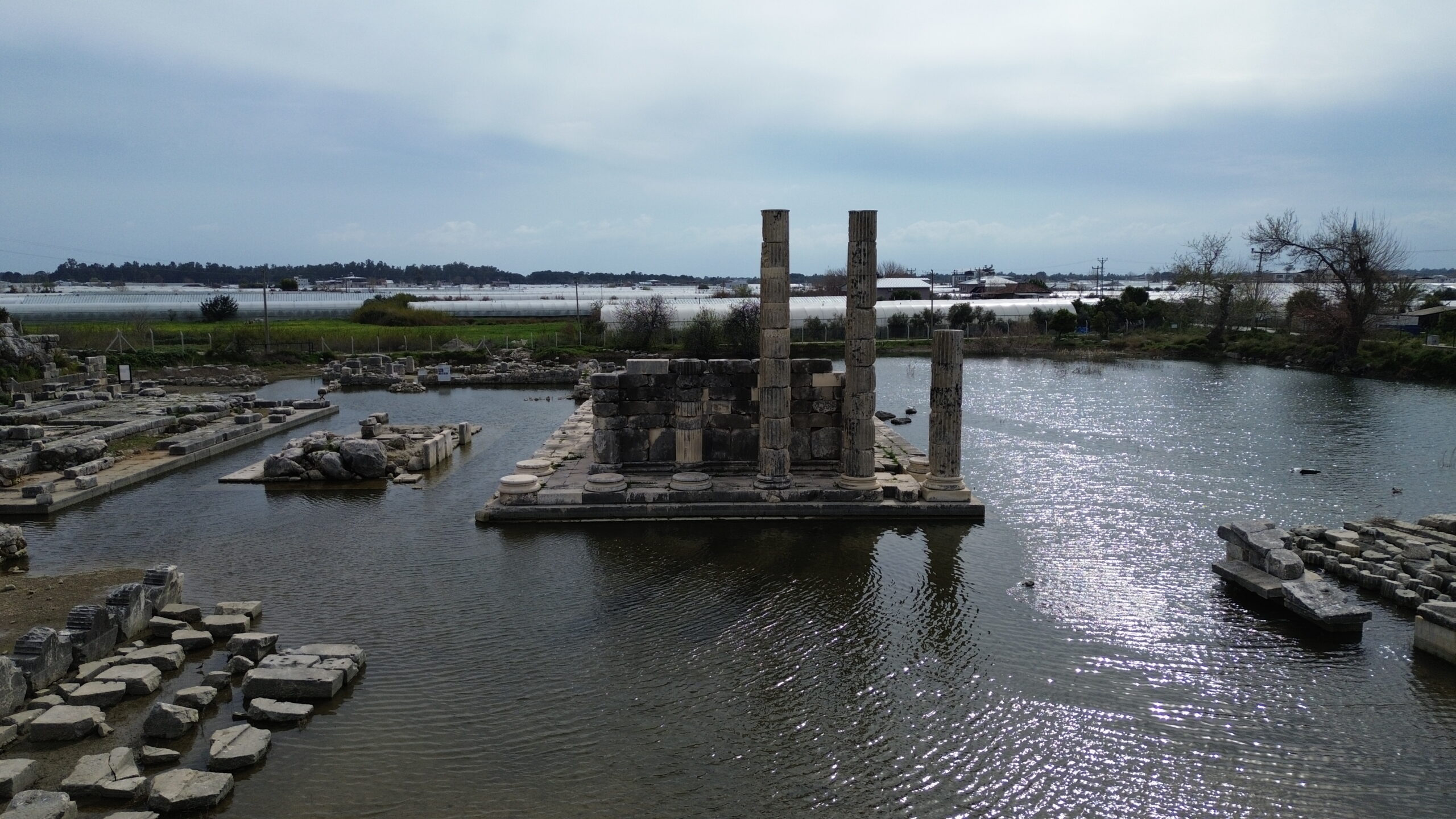 The monumental Temple of Leto in the Ionic order, partially submerged under water during winter months in Letoon Ancient City, with the ancient theater steps rising in the background.